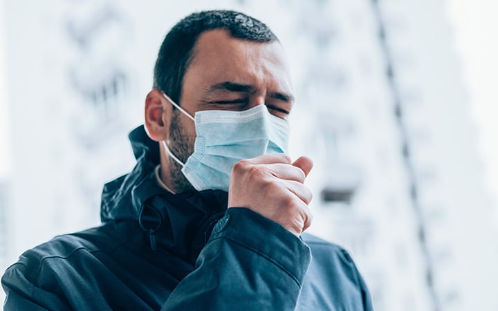 A young man with closed eyes wearing a face protective mask and coughing in his arm